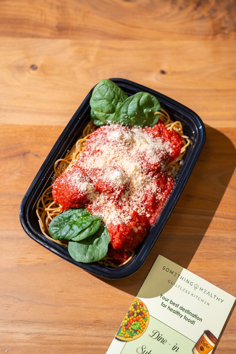 Spaghetti with red sauce, meatballs, parmesan, and spinach in a black to-go container next to a Something Healthy menu.
