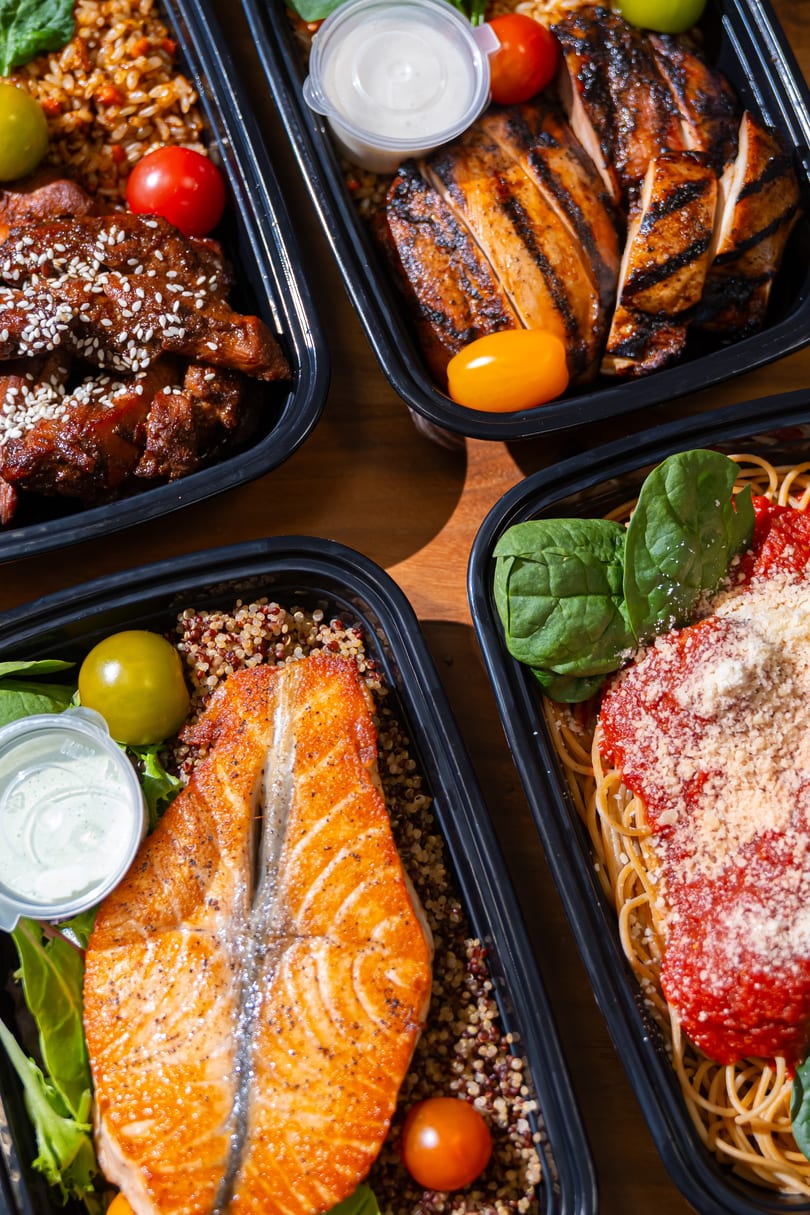 Four meal prep containers with grilled chicken, salmon, pasta, and sesame chicken, each with sides.