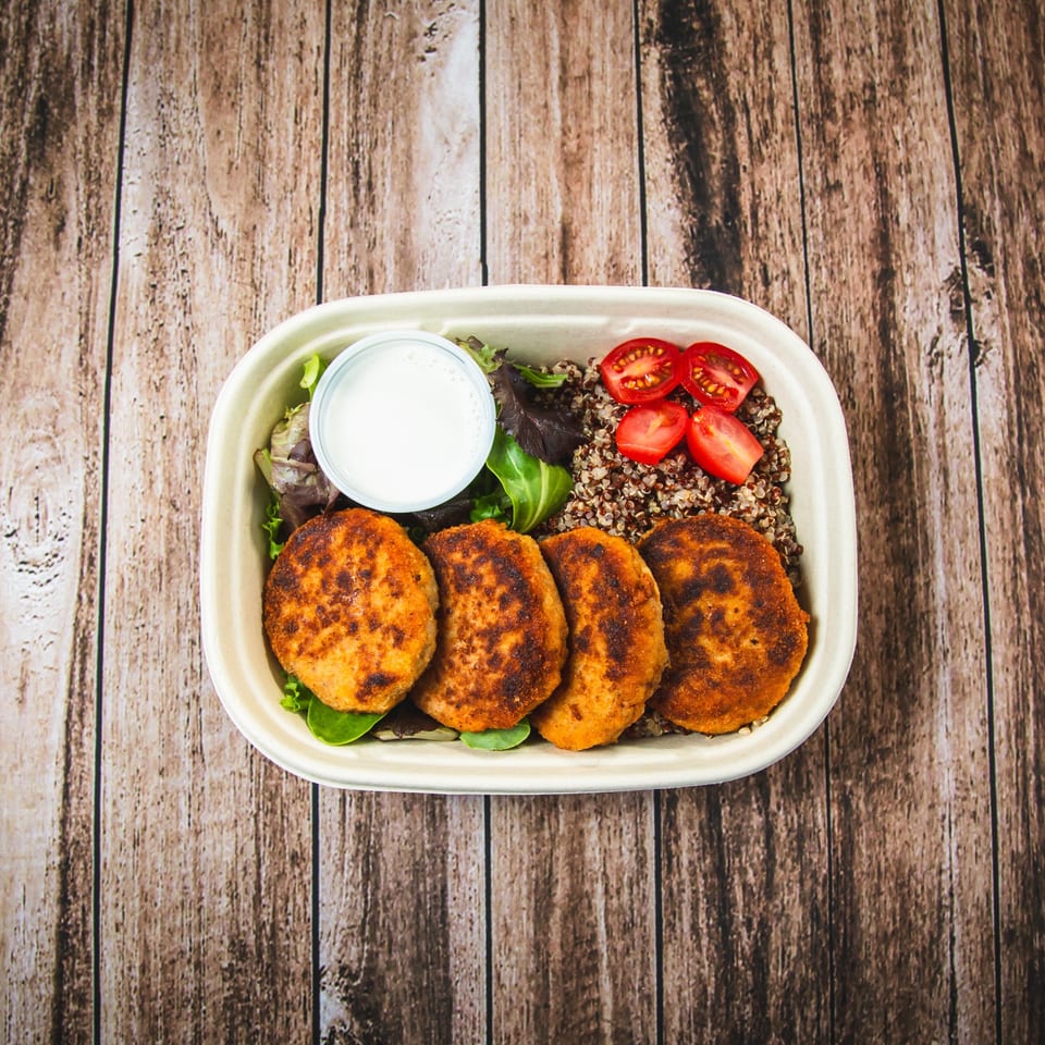 A meal prep container with four crispy patties, quinoa, cherry tomatoes, salad, and dressing.