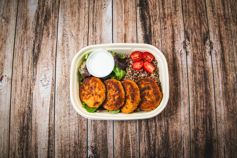 A meal prep container with four crispy patties, quinoa, cherry tomatoes, salad, and dressing.