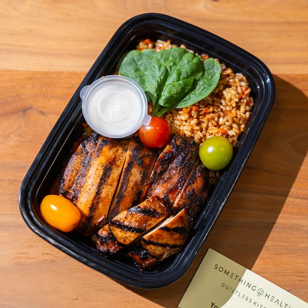 Grilled chicken, rice, spinach, cherry tomatoes, and sauce in a black to-go container.