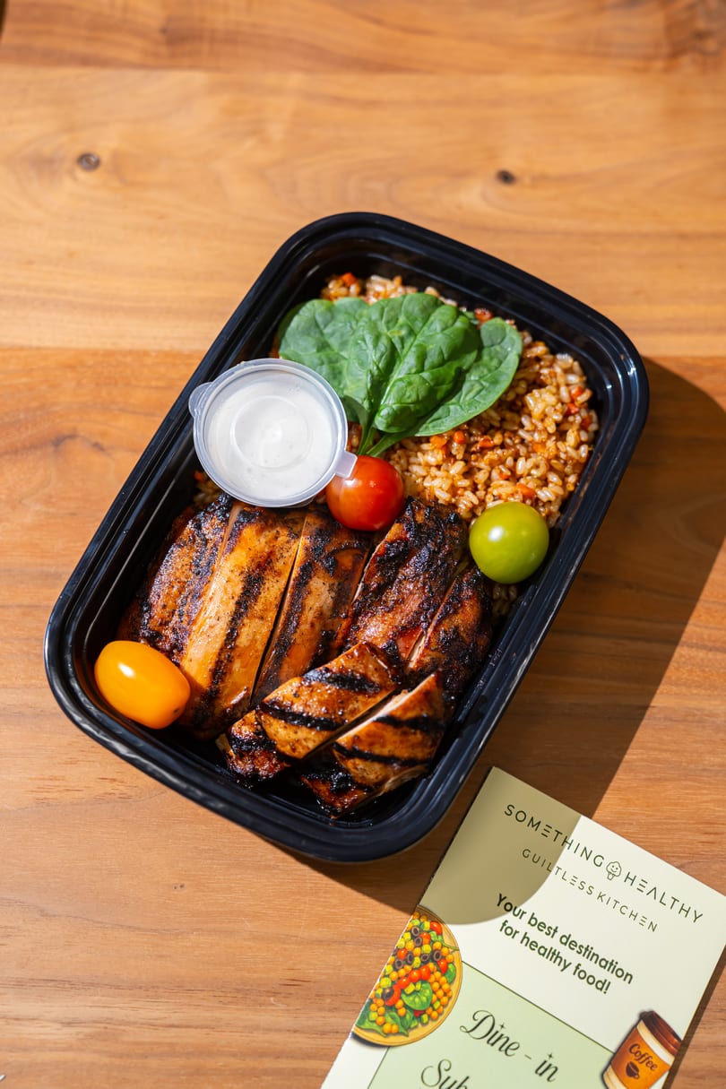 Grilled chicken, rice, spinach, cherry tomatoes, and sauce in a black to-go container.