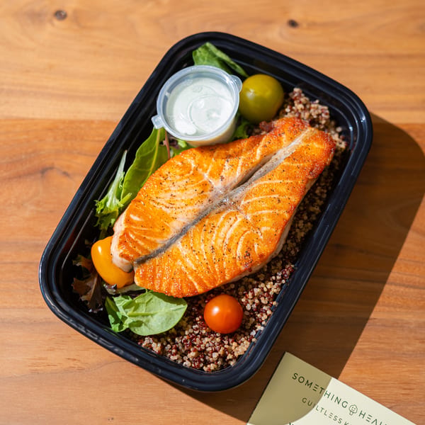 Something Healthy's salmon and quinoa meal with salad and dressing, next to a menu on a wooden table.