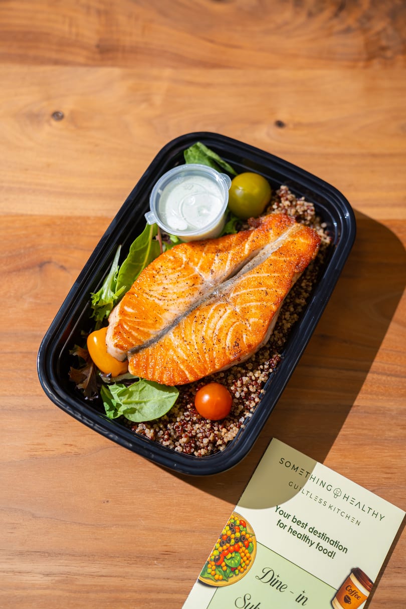 Something Healthy's salmon and quinoa meal with salad and dressing, next to a menu on a wooden table.