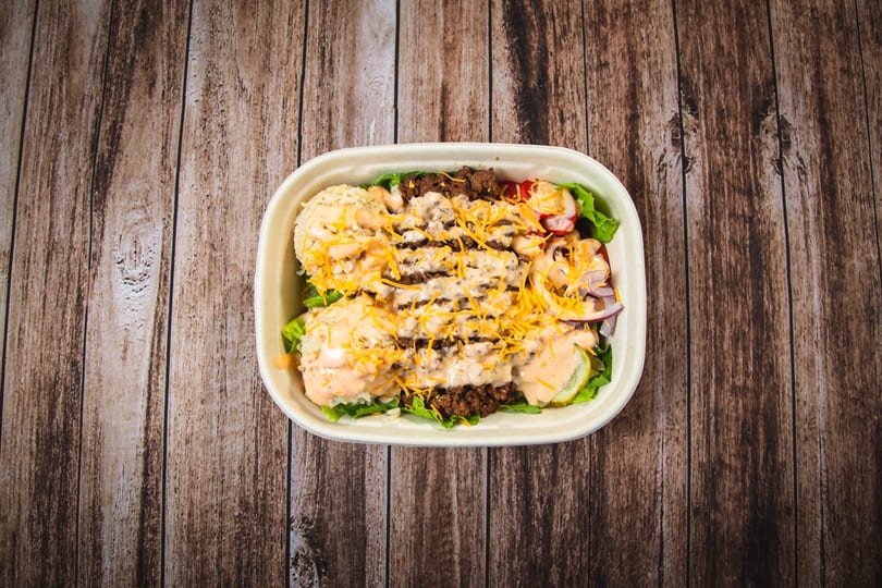 Overhead view of a burger bowl with ground beef, rice, lettuce, cheese, and sauce on wood.