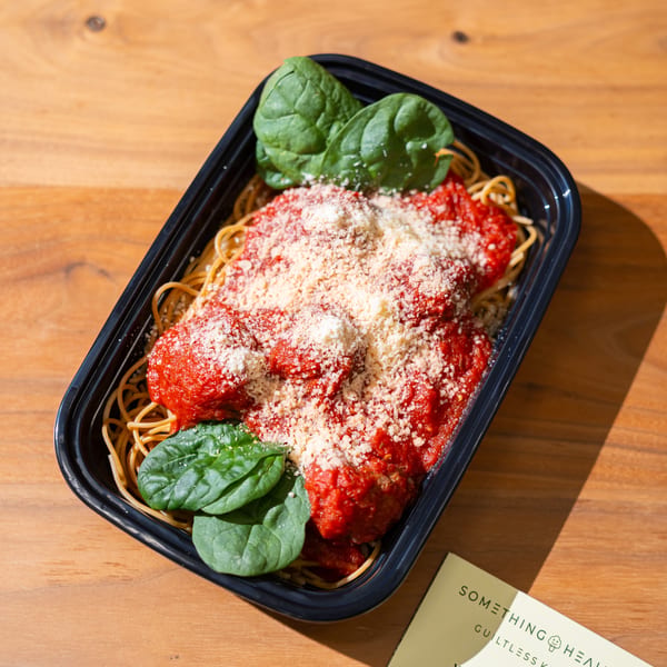 Spaghetti with meatballs, red sauce, parmesan, and spinach in a black to-go container next to a Something Healthy menu.