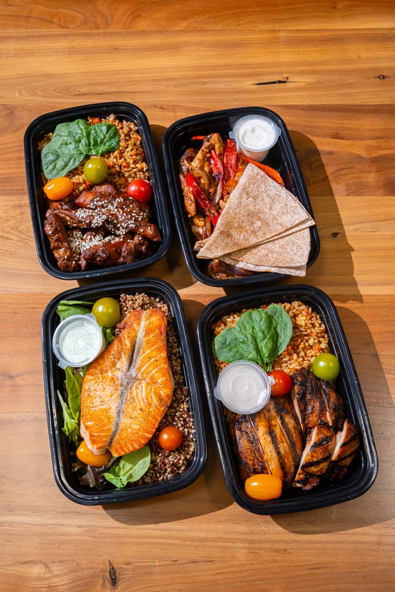 Four meal prep containers with salmon, chicken, fajitas, and sesame chicken on a wooden table.